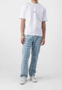 T-shirt bianco con logo blu, pantaloni in denim azzurro chiaro con motivo ripetuto "HUGO", e sneaker bianche. Outfit casual.