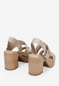 Sandali con tacco quadrato in pelle beige, dotati di cinturini incrociati e un grande dettaglio a fibbia. Tacco in legno con una superficie liscia e forma arrotondata.