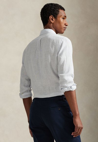 Ανοιχτό μπλε πουκάμισο με ρίγες και μανίκια που έχουν γυριστεί, με ορατή υφή υφάσματος. Φορεμένο με σκούρο παντελόνι, παρουσιάζοντας ένα ραμμένο πίσω σχέδιο και λεπτομέρειες με κουμπιά.