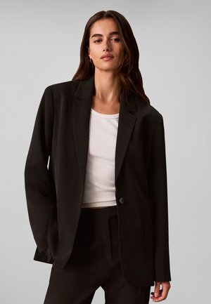Kvinde med langt brunt hår iført en sort oversized blazer og hvid top, stående mod en ensfarvet lys grå baggrund.