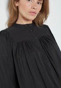 Must kaaelusega must blouse, nupuga kinnitamine ja vertikaalsed tekstuuriribad; omab pikki varrukaid ja kogutud kangast volüümi tekitamiseks.
