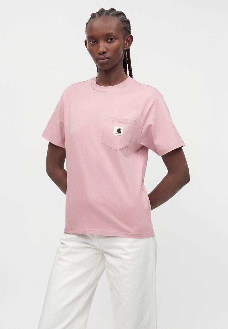 Woman with braided hair wearing a light pink short-sleeve T-shirt with a chest pocket logo and white pants against a plain background.