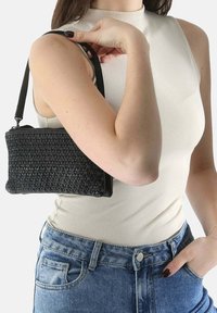 Borsa a tracolla nera intrecciata con un design rettangolare e liscio e una singola tracolla regolabile, tenuta contro una maglietta neutra e jeans. 