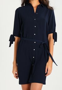 Námořnická modrá šaty košile vyrobené z měkké látky, s ozdobným knoflíkovým zapínáním vpředu, zavázanými rukávy a pásem v pase. Hladká textura s minimálními detaily.