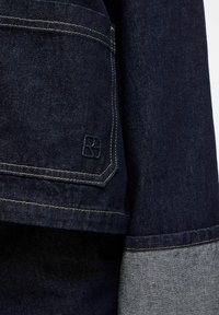 Donkerblauwe denim jacketmouw met opgerolde manchet en een klein geborduurd logo nabij de zoom met zichtbare oranje stiksel.