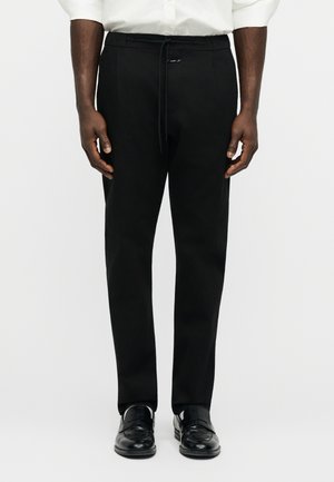 Muž oblečený v čiernych na mieru šitých nohaviciach sťahovaných šnúrkou v páse, čierne spoločenské topánky a biela košeľa, stojaci pred jednoduchým bielym pozadím.