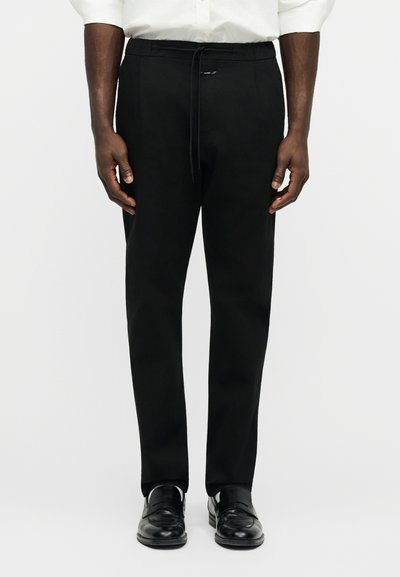 Homme portant un pantalon noir sur mesure avec taille à cordon, chaussures habillées noires et chemise blanche, debout devant un fond blanc uni.