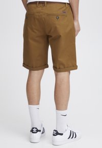 Brown cotton shorts with rolled hems, featuring two back pockets with buttons, worn with white socks and black-and-white sneakers.