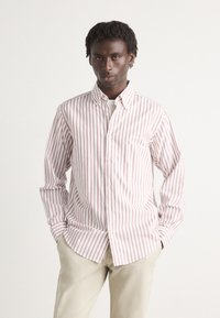 Camicia bianca con sottili righe rosse verticali, colletto button-down, maniche lunghe e orlo curvo. Indossata con pantaloni grigi chiari.
