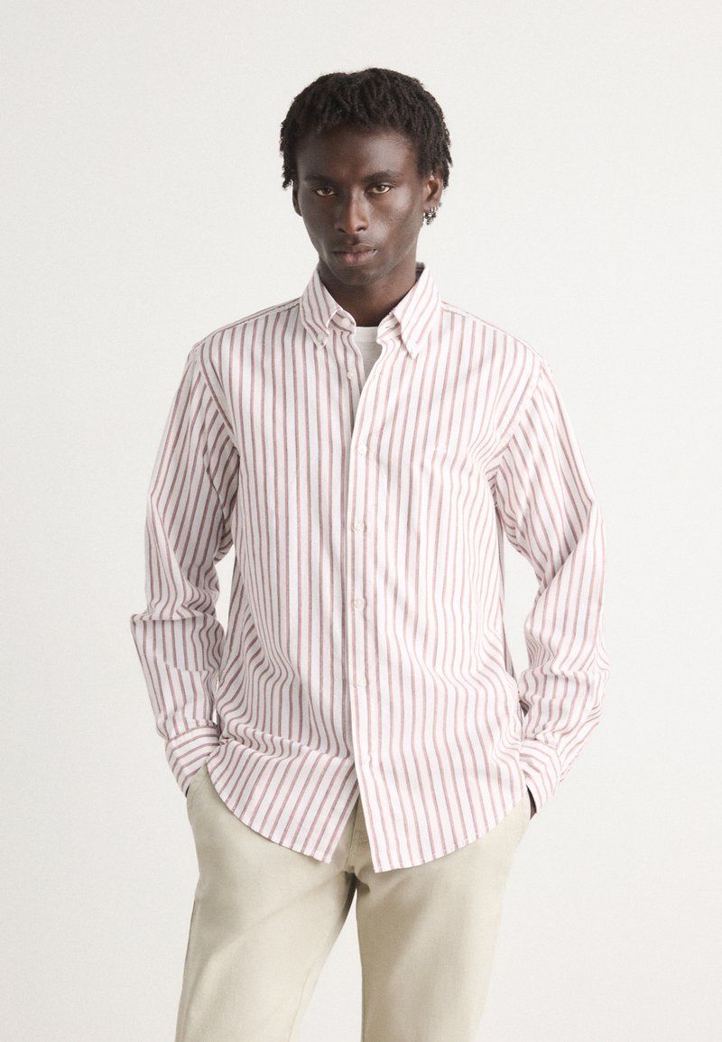 Camicia bianca con sottili righe rosse verticali, colletto button-down, maniche lunghe e orlo curvo. Indossata con pantaloni grigi chiari.