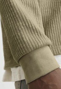 Olivgrüner, gerippter Strickpulloverärmel mit einem glatten, enganliegenden Bündchen. Die Textur ist weich und verfügt über eine kontrastierende helle Innenlage.