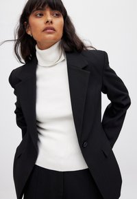 Juodas švarkas su struktūrizuotu dizainu, vienu užsegimu ir išpjautomis atlapomis, vilkimas ant raukšlėto balto megztinio su aukštu kaklu.