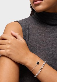 Pulsera de cadena de plata con un acento negro en una muñeca. El tono de piel es medio, lleva una parte superior sin mangas en gris oscuro con un escote alto.