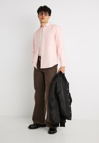 Jeune homme portant des lunettes, chemise rose à boutons, pantalon marron, chaussures noires, tenant une veste noire dans un studio blanc.