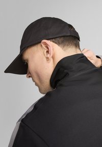 Gorra negra con visera curvada, combinada con una chaqueta negra de cuello alto que presenta detalles en gris. Textura de tela suave, diseño minimalista.