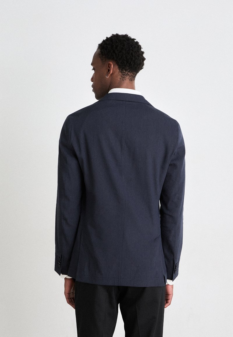 Veste de costume marine avec une coupe ajustée, revers crantés, fente simple et poignets boutonnés. Le tissu est lisse avec une texture subtile.