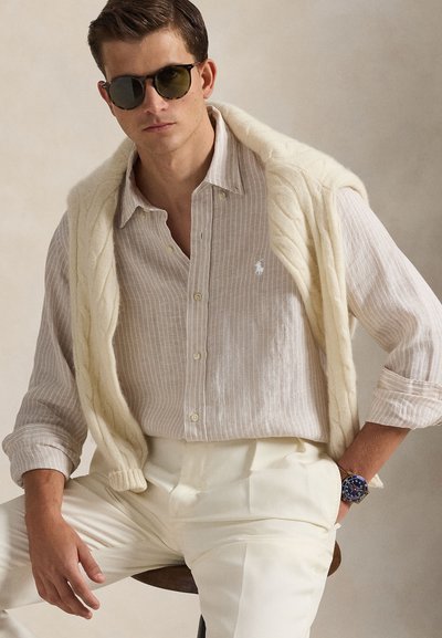 Homme portant des lunettes de soleil foncées, une chemise à rayures claires boutonnée, un pull en tricot crème drapé sur les épaules, un pantalon blanc et une montre bleue au poignet, assis sur un tabouret.