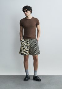 Uomo che indossa una maglietta aderente marrone, occhiali da sole neri, pantaloncini con motivi a stampa animale e metà a scacchi, calzini grigi e scarpe nere.