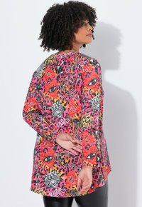 T-shirt à manches longues avec un motif imprimé animal multicolore, présentant des fleurs rouges, des images de tigres et du texte écrit à la main sur un tissu texturé.