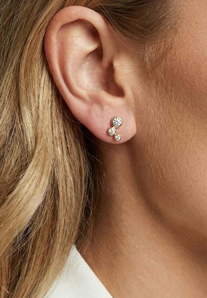 Øre med tre runde ørestikker med klare ædelstene arrangeret i en nedadgående trekant, båret på lys hud med synligt blondt hår.