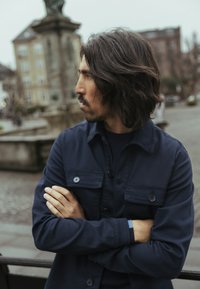 Tumši zils pogājams žakete ar divām krūšu kabatām un brīvu piegriezumu, kopā ar atbilstošu apaļu kakla kreklu. Fakturēts audums un gari mati.