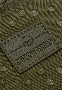 Dunkelolivgrüner Stoff mit einer strukturierten Oberfläche, ausgestattet mit einem geprägten Logo-Patch und klaren, runden Nieten. Robustes und wasserabweisendes Design.