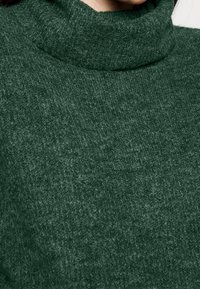 Gros plan d'une personne portant un pull à col roulé vert foncé en maille pelucheuse avec un tissu doux et texturé.