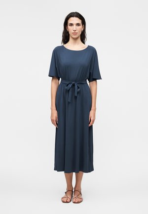 Kobieta stoi przodem, ubrana w granatową sukienkę midi z krótkimi rękawami i związanym paskiem w talii, w połączeniu z brązowymi, płaskimi sandałami.