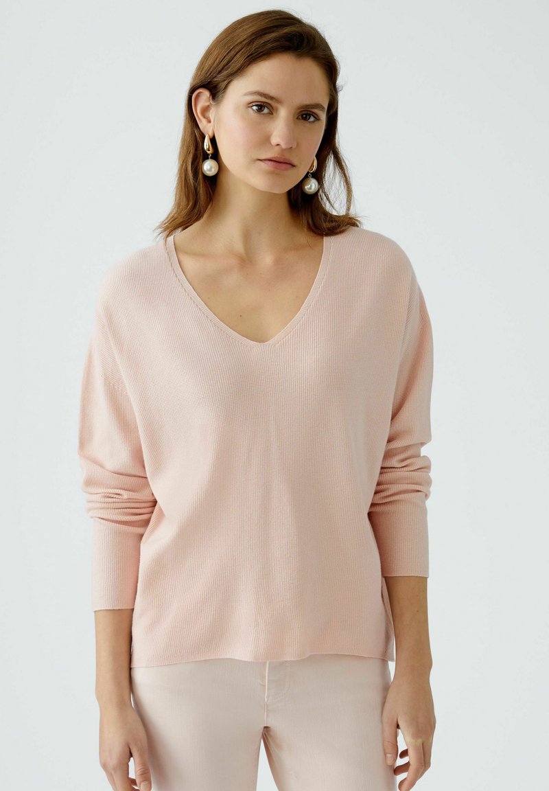 Light pink, ribbed knit sweater with a V-neck and long sleeves. Fabric appears soft with a relaxed fit, styled with matching light pants.