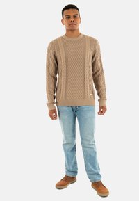 Maglione beige a maglia con motivi testurizzati, collo rotondo e polsini a costine, abbinato a jeans azzurri chiari e scarpe marroni.