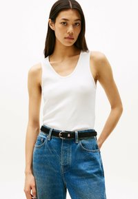 Weißes, geripptes Trägershirt kombiniert mit hochtaillierten blauen Jeans und einem schwarzen, taillierten Gürtel. Das Trägershirt hat einen runden Ausschnitt und sitzt figurbetont.