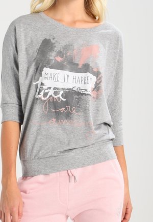 Femme portant un sweat-shirt gris avec l'inscription « MAKE IT HAPPEN » en texte rose cursif, associée à un pantalon rose clair avec un cordon de serrage.