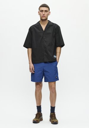 Black short-sleeve button-up shirt with two front pockets, paired with blue shorts featuring a logo patch. Brown and black hiking shoes.