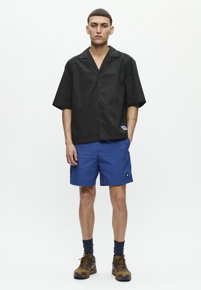 Black short-sleeve button-up shirt with two front pockets, paired with blue shorts featuring a logo patch. Brown and black hiking shoes.