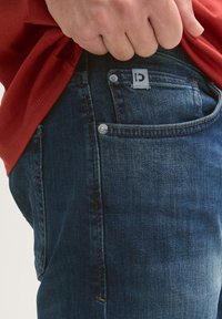 Sötétkék farmernadrág struktúrált felülettel, ezüstösen csillogó fémalkatrészekkel és márkajelzéssel a derékrészen, egy piros inggel viselve.