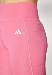Ružové legíny s vysokým pásom vyrobené z elastického materiálu, s bočnou vreckom a nenápadným detailom šitia. Biele logo značky na stehne.