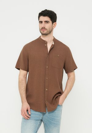 Άνδρας με κοντά σκούρα μαλλιά που φοράει καφέ κοντομάνικο πουκάμισο με κουμπιά και τσέπη στο στήθος και ανοιχτόχρωμο μπλε τζιν, όρθιος μπροστά σε λευκό φόντο.