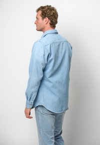 Camisa de mezclilla azul clara, de manga larga, con puños abotonados, dobladillo curvado, diseño de yugo en la parte trasera, combinada con jeans azul claro a juego.