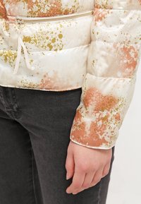Prošívaná bunda s krémovým základem a abstraktními růžovo-zlatými skvrnitými vzory. Obsahuje kapsu na zip a elastické manžety.