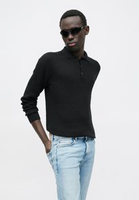 Polo preta em malha com gola, mangas compridas e pala com botões, combinada com jeans azul claro. O corte é descontraído.