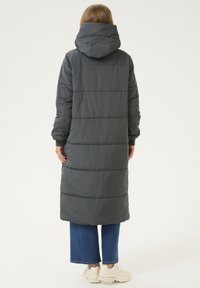Manteau rembourré long couleur gris foncé, doté d'une capuche et d'un matelassage horizontal. Associé à un jean bleu à jambes larges et des baskets beiges.