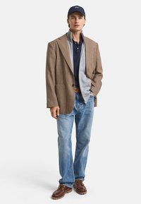 Blazer marrón a cuadros, sweater gris con cremallera, polo azul marino, jeans azul claro y zapatos marrones. Conjunto casual en capas con texturas y colores mezclados.
