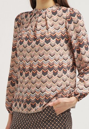 Kvinde iført en langærmet bluse med et flerfarvet chevronmønster og matchende mønstrede bukser, hånd hvilende i lommen.