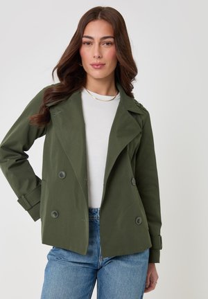 Vrouw met lang bruin haar draagt een olijfgroene dubbelgeknoopte jas over een wit overhemd en blauwe spijkerbroek, poserend tegen een effen lichte achtergrond.