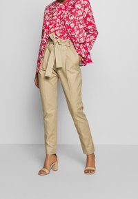 Roze bloemenprint jack met witte accenten, gecombineerd met beige high-waisted broek met een stoffen riem en een zachte textuur. Heldere blokhakken.