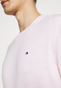 Rosa stickad tröja med ribbad krage, prydd med en liten broderad logotyp i rött, vitt och blått på vänster bröst.