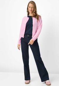 Cardigan rosa con bottoni, indossato sopra una maglia blu scuro, abbinato a pantaloni svasati in denim scuro e sneakers rosa chiaro. Tessuto liscio.