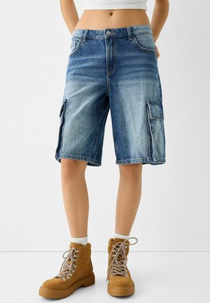 Person trägt ausgewaschene blaue Denim-Cargoshorts, weiße Socken und braune Schnürstiefel und steht vor einem weißen Hintergrund.