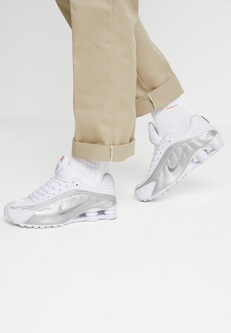 Personne portant un pantalon beige et des baskets blanches Nike avec des accents argentés sur un fond blanc uni.