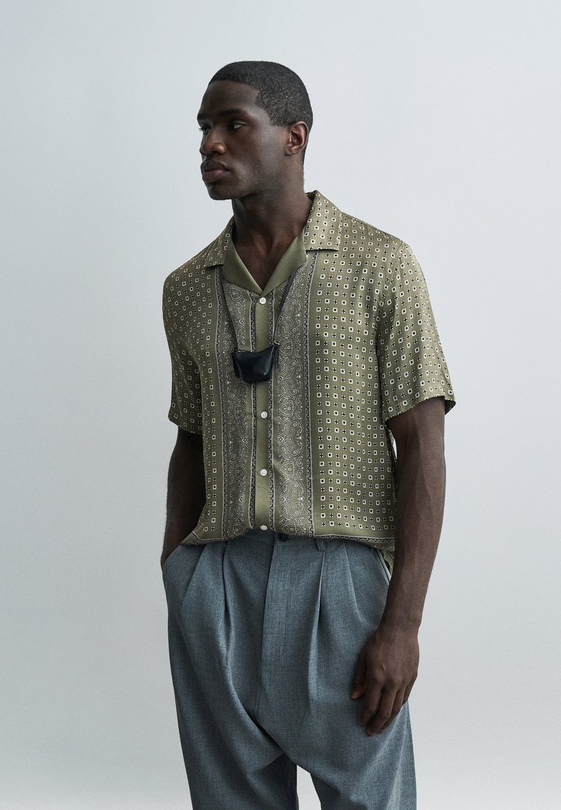 Άνδρας που φοράει πράσινο πουκάμισο με σχέδια και κοντά μανίκια, γκρι πιετέ παντελόνι και μαύρη μάσκα προσώπου κρεμασμένη γύρω από το λαιμό, στέκεται μπροστά σε απλό φόντο.